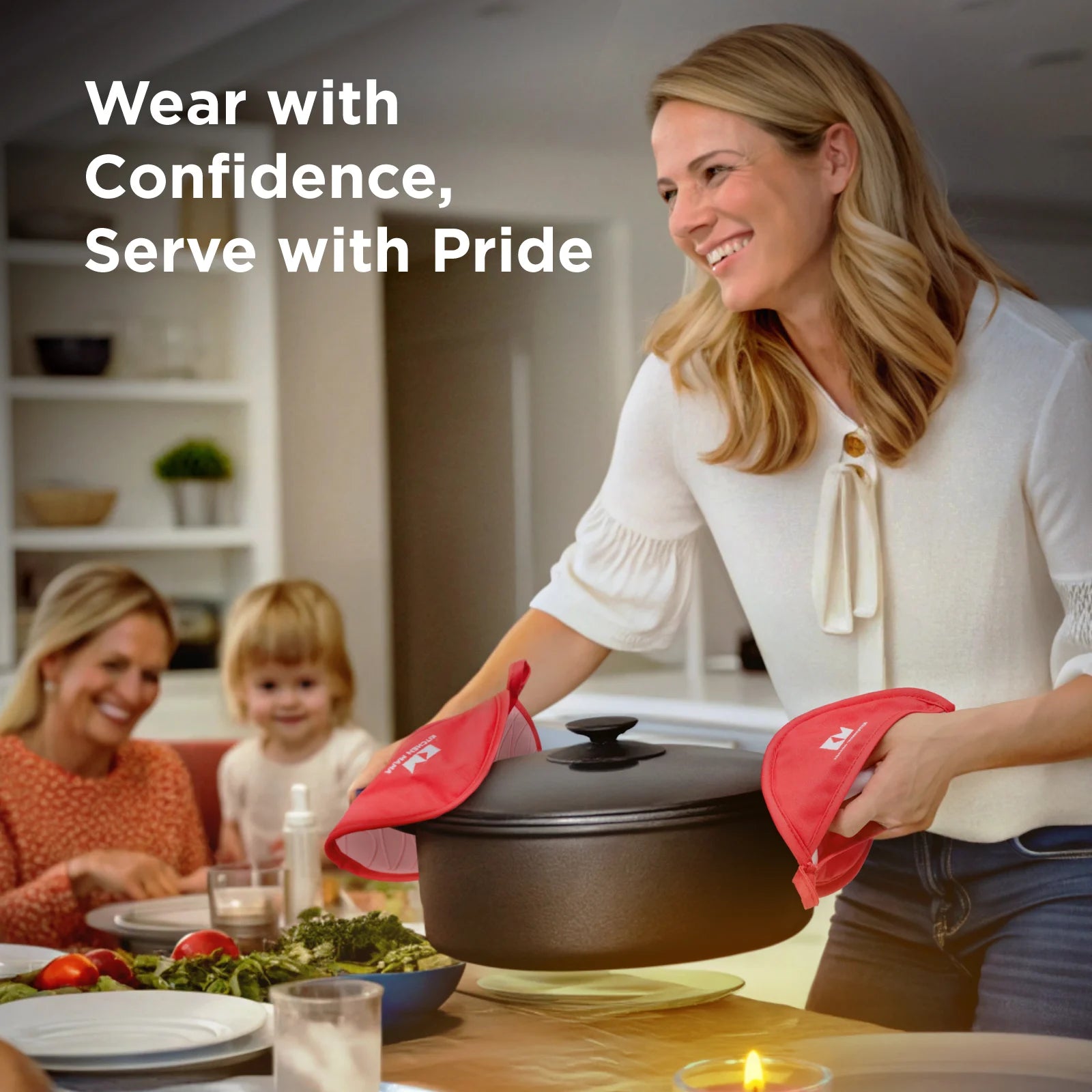 A smiling woman wearing white serves a hot meal using red silicone pot holders, with a cast iron pot in hand, as a child and another woman smile at the dining table in a warmly lit kitchen.